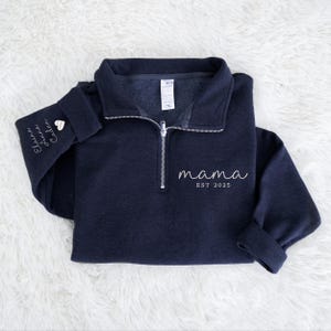 May include: A navy blue pullover sweatshirt with a quarter-zip neckline. The word "mama" and "EST 2025" are printed in white script on the front. The left sleeve has embroidered names and a heart. The sweatshirt is folded on a white, fluffy surface.