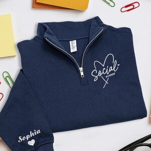 May include: A navy blue quarter-zip sweatshirt with the words "Social Worker" in white script and a heart design. The name "Sophia" is printed on the sleeve with a heart. The sweatshirt has a silver zipper.