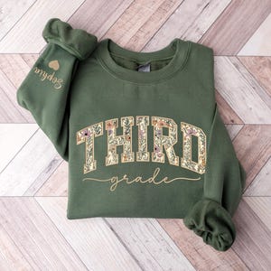 May include: Green sweatshirt featuring the words "THIRD grade" in floral lettering. The word "grade" is in cursive. The left sleeve has the name "Sophia" embroidered with a small gold heart. The sweatshirt is folded on a wood-effect surface.