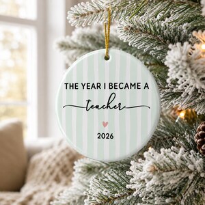 May include: A round white ceramic ornament featuring vertical light green stripes. The text reads THE YEAR I BECAME A teacher with a small pink heart and the year 2026 printed below. A gold string is attached to the top for hanging on a tree.