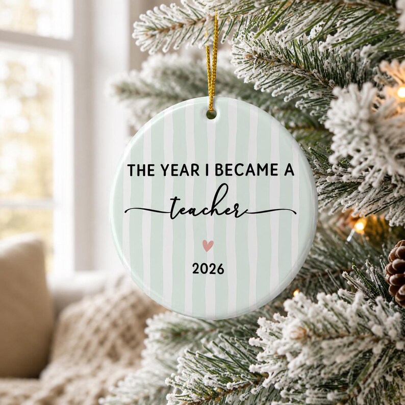 May include: A round white ceramic ornament featuring vertical light green stripes. The text reads THE YEAR I BECAME A teacher with a small pink heart and the year 2026 printed below. A gold string is attached to the top for hanging on a tree.