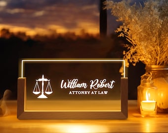 May include: A wooden-framed LED light with the text "William Robert ATTORNEY AT LAW" and a scales of justice symbol. The light is on, illuminating the text and symbol in a warm glow. The background features a sunset and decorative elements.
