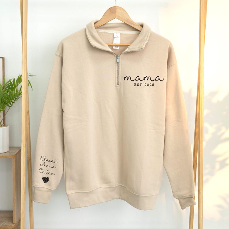 May include: A beige pullover sweatshirt with a quarter-zip neckline. The word "mama" and "EST 2025" are printed in black script on the front. The left sleeve has the names Elaina, Anna, and Caden, with a heart symbol below.