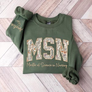 May include: An olive green sweatshirt with the letters "MSN" in a floral design. Below the letters is the text "Master of Science in Nursing". The sleeve has the name "Amelia" embroidered in gold.