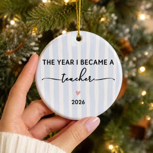 May include: A round white ceramic ornament featuring vertical light blue stripes. The text reads THE YEAR I BECAME A teacher with a small pink heart and the year 2026 printed below. A gold string is attached to the top for hanging purposes.