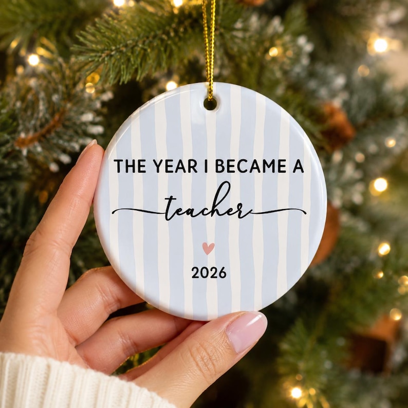 May include: A round white ceramic ornament featuring vertical light blue stripes. The text reads THE YEAR I BECAME A teacher with a small pink heart and the year 2026 printed below. A gold string is attached to the top for hanging purposes.