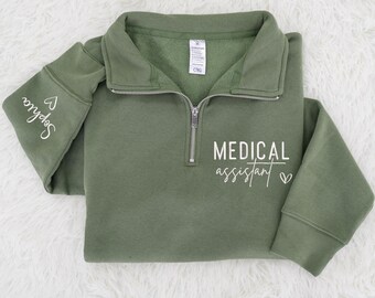 May include: Olive green quarter-zip sweatshirt with the words "MEDICAL assistant" in white script. The left sleeve has the name "Sophia" in white script. The sweatshirt has a silver zipper and a folded collar.