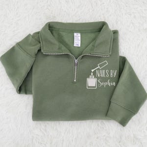May include: An olive green quarter-zip sweatshirt featuring the text "NAILS BY Sophia" and a white nail polish bottle graphic. The sweatshirt has a collared neckline, long sleeves, and a silver zipper.
