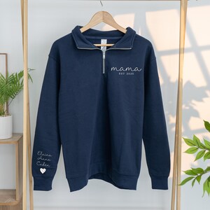 May include: Navy blue sweatshirt with a quarter-zip neckline. The word "mama" and "EST 2025" are printed on the front. The left sleeve has the names "Elaina, Anna, Caden" and a heart symbol.
