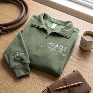 May include: An olive green quarter-zip sweatshirt featuring white embroidered text on the chest that reads "PILATES instructor" with a small heart. The sleeve is embroidered with the name "Sophia" and a heart. The collar has a silver zipper.