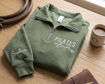 May include: An olive green quarter-zip sweatshirt featuring white embroidered text on the chest that reads "PILATES instructor" with a small heart. The sleeve is embroidered with the name "Sophia" and a heart. The collar has a silver zipper.