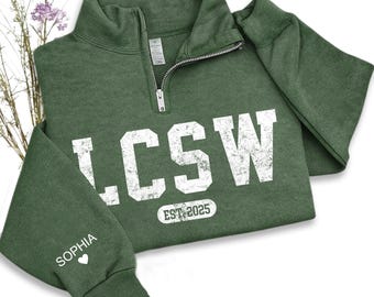 May include: A green quarter-zip sweatshirt with the letters LCSW in white, distressed font. The text "EST. 2025" is below the letters. The sleeve cuff has the name "Sophia" and a heart symbol. The sweatshirt is made of a soft material.