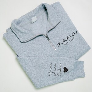 May include: A light gray fleece pullover with a silver zipper and the word "mama" printed in black script, along with "EST 2025". The sleeve features the names Elaina, Anna, and Caden, and a black heart.
