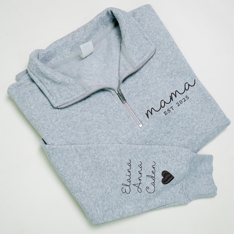 May include: A light gray fleece pullover with a silver zipper and the word "mama" printed in black script, along with "EST 2025". The sleeve features the names Elaina, Anna, and Caden, and a black heart.