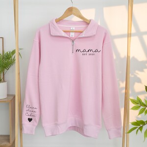 May include: A light pink sweatshirt with a quarter-zip collar. The word "mama" and "EST 2025" are printed in black on the chest. The left sleeve has the names "Elaina, Anna, Caden" and a heart symbol printed in black. The sweatshirt is hanging on a wooden hanger.