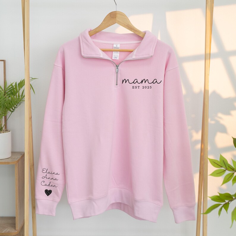 May include: A light pink sweatshirt with a quarter-zip collar. The word "mama" and "EST 2025" are printed in black on the chest. The left sleeve has the names "Elaina, Anna, Caden" and a heart symbol printed in black. The sweatshirt is hanging on a wooden hanger.