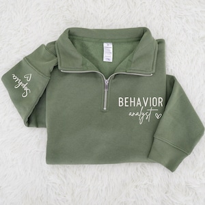 May include: Olive green quarter-zip sweatshirt with the words "BEHAVIOR analyst" in white script. The sleeve has the name "Sophia" in white script. The sweatshirt has a silver zipper and a folded collar.