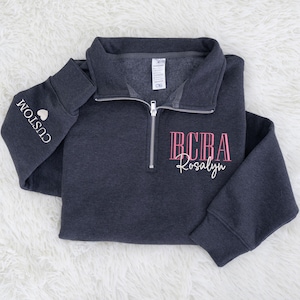 May include: A dark gray quarter-zip sweatshirt with the text "BCBA Rosalyn" in pink script on the front. The left sleeve has the word "CUSTOM" and a heart. The sweatshirt is made of a soft material.