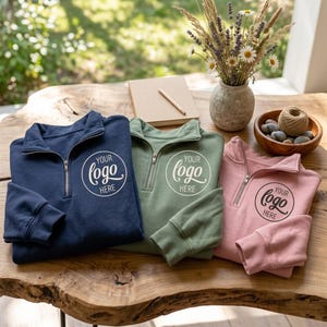 Peut inclure: Trois sweat-shirts à fermeture éclair quart de longueur en bleu marine, vert sauge et rose poudré, chacun arborant un motif circulaire sur la poitrine avec le texte "YOUR LOGO HERE". Les vêtements sont disposés sur une surface en bois.
