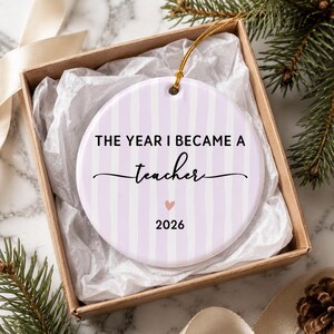 May include: A round ceramic ornament with a light purple and white vertical striped pattern. The text reads THE YEAR I BECAME A teacher with a small pink heart and the year 2026 printed below. A gold string is attached for hanging purposes.