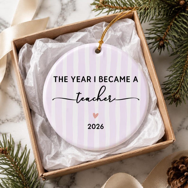May include: A round ceramic ornament with a light purple and white vertical striped pattern. The text reads THE YEAR I BECAME A teacher with a small pink heart and the year 2026 printed below. A gold string is attached for hanging purposes.