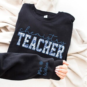 May include: Black sweatshirt featuring the words "chemistry TEACHER" in blue script and a blue and white floral design. The sleeve displays "Mrs. Wilson" and a science symbol in blue. The sweatshirt is folded on a white surface.
