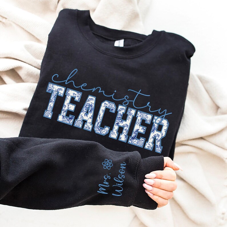 May include: Black sweatshirt featuring the words "chemistry TEACHER" in blue script and a blue and white floral design. The sleeve displays "Mrs. Wilson" and a science symbol in blue. The sweatshirt is folded on a white surface.