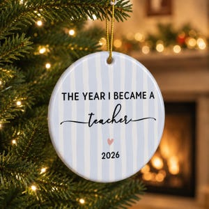 May include: A round white ceramic ornament featuring vertical light blue stripes. The text reads THE YEAR I BECAME A teacher with a small pink heart and the year 2026 printed below. A gold string is attached to the top for hanging on a tree.