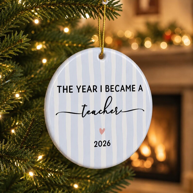 May include: A round white ceramic ornament featuring vertical light blue stripes. The text reads THE YEAR I BECAME A teacher with a small pink heart and the year 2026 printed below. A gold string is attached to the top for hanging on a tree.