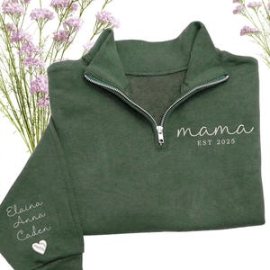May include: A dark green sweatshirt with a quarter-zip neckline. The word "mama" and "EST 2025" are embroidered in white on the front. The left sleeve has the names "Elaina, Anna, Caden" embroidered in white, along with a white heart.