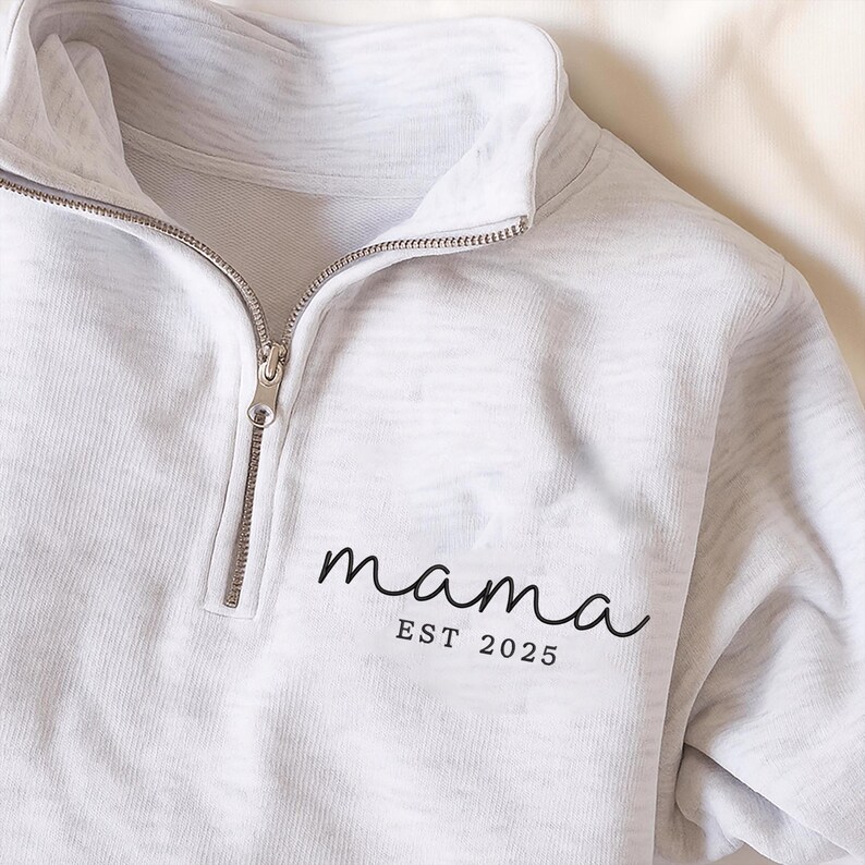 May include: A light gray pullover sweatshirt with a quarter-zip neckline. The word "mama" is printed in a cursive font, with "EST 2025" below it. The sweatshirt has a ribbed texture and a silver zipper.