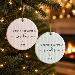 May include: Two round ceramic ornaments hanging by gold strings. One is light pink with white vertical stripes, the other is light green with white vertical stripes. Both feature the black text THE YEAR I BECAME A teacher, a small pink heart, and 2026.