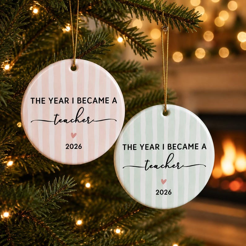 May include: Two round ceramic ornaments hanging by gold strings. One is light pink with white vertical stripes, the other is light green with white vertical stripes. Both feature the black text THE YEAR I BECAME A teacher, a small pink heart, and 2026.