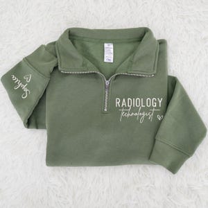 May include: Olive green quarter-zip sweatshirt with the text "Radiology Technologist" in white script. The left sleeve has the name "Sophia" and a heart symbol. The sweatshirt has a folded collar and a silver zipper.