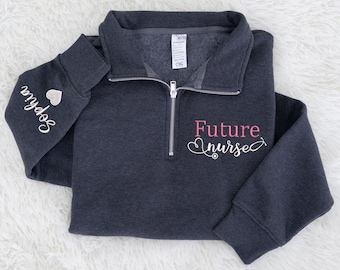 Könnte beinhalten: Ein anthrazitfarbenes Sweatshirt mit Viertelreißverschluss und dem Schriftzug "Future nurse" in Rosa und Weiß. Auf dem linken Ärmel ist der Name "Sophia" mit einem kleinen Herz gestickt. Das Sweatshirt ist ordentlich gefaltet.