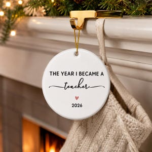 May include: A round white ceramic ornament hanging from a gold metal hook. The text reads THE YEAR I BECAME A teacher in black, with a small pink heart and the year 2026 printed below. The ornament is suspended by a thin gold string.