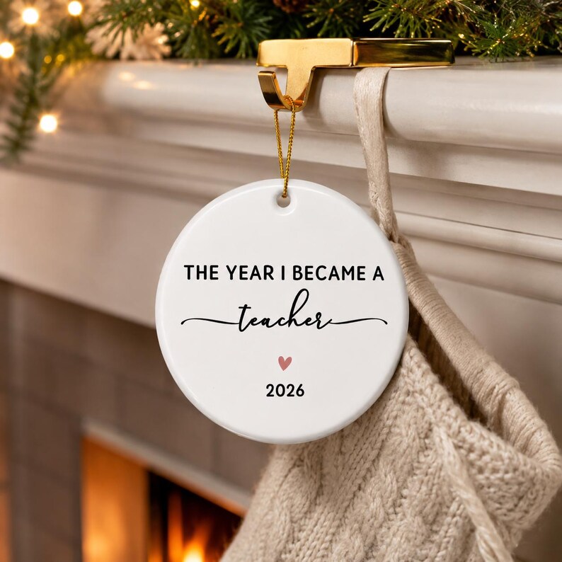 May include: A round white ceramic ornament hanging from a gold metal hook. The text reads THE YEAR I BECAME A teacher in black, with a small pink heart and the year 2026 printed below. The ornament is suspended by a thin gold string.
