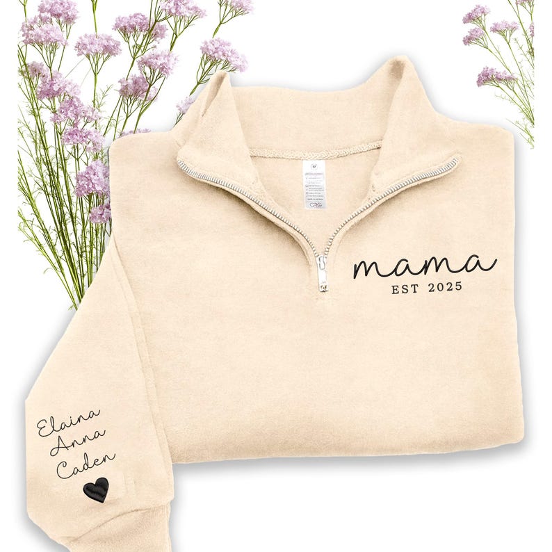 May include: Cream-colored fleece pullover with a quarter-zip neckline. The word "mama" and "EST 2025" are printed in black script on the front. The left sleeve has the names "Elaina Anna Caden" and a black heart.