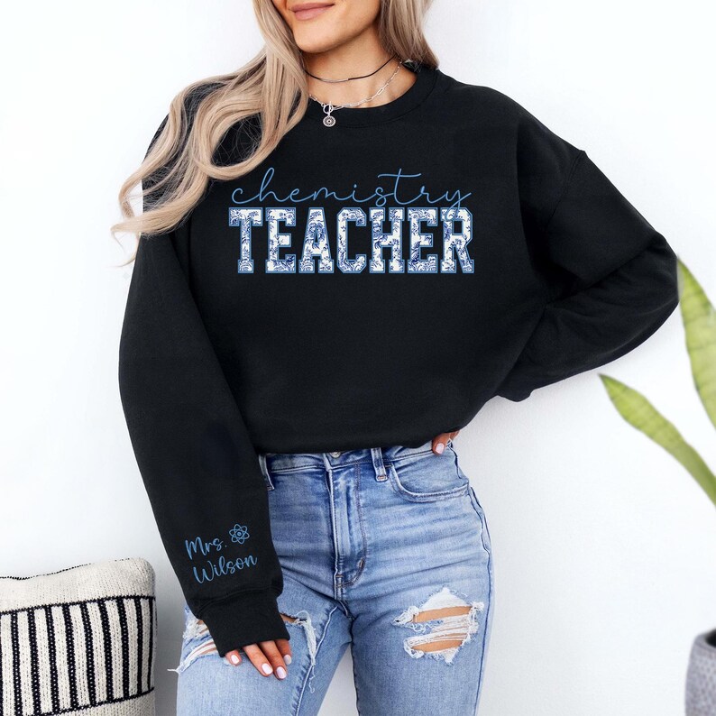 May include: Black sweatshirt featuring the words "chemistry TEACHER" in a blue floral design. The sleeve displays "Mrs. Wilson" and a blue atom symbol. The sweatshirt is worn with blue jeans.