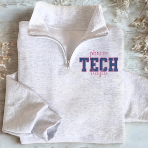 May include: A light gray quarter-zip sweatshirt with the text "pharm TECH Kayla" in pink and blue lettering. The sweatshirt has a high collar and a silver zipper. The sleeves are long and cuffed.