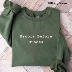 May include: A military green sweatshirt with the text "Proofs Before Grades" in white. The crew neck sweatshirt has long sleeves and is folded neatly. The background is a light brown wood grain pattern.