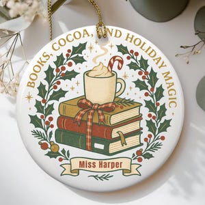 Pode incluir: Enfeite de Natal em cerâmica branca com fio dourado. O enfeite apresenta uma pilha de livros, uma caneca de cacau com uma bengala doce e o texto "BOOKS, COCOA, AND HOLIDAY MAGIC". Uma faixa diz "Miss Harper".