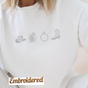 Könnte beinhalten: Weißes Sweatshirt mit gestickten Designs eines Cowboyhuts, eines Cocktails, eines Rings und eines Cowboy-Stiefels. Das Wort "Embroidered" ist unten in brauner Schrift gedruckt.