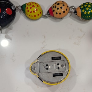 May include: A row of four colorful metal ladybug toys connected by metal links, featuring black, yellow, tan, and green bodies with polka dots. Below the row, a single yellow ladybug toy is positioned upside down, revealing its metal base and wheels.