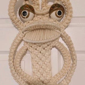May include: A cream-colored macrame wall hanging shaped like a frog, featuring two large ceramic eyes with brown and green speckled lids and black pupils. The piece is constructed from knotted cord and is suspended from a curved bamboo hoop at the top.