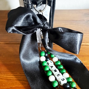Op de afbeelding: Een doorzichtige tas met ritssluiting met een zwarte satijnen strik en een kralen sleutelhanger. De sleutelhanger spelt "HAWKS" met witte letterkralen, groene en zwarte kralen. De strik is om een zilveren ring gebonden.