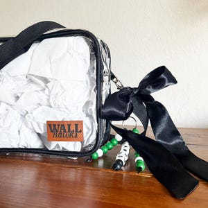 May include: A clear, square bag with a black strap and the text "WALL hawks" on a brown patch. The bag is filled with white paper and has a black bow and beaded keychain attached.