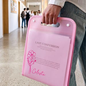 May include: A translucent pink plastic document folder with a built-in handle and two white snap closures. The front features a printed floral line drawing, the text "LAST CONFESSION" in a serif font, and the name "Celeste" in a script font.