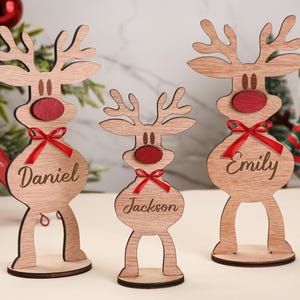May include: Wooden reindeer Christmas decorations with red noses and bows. Each reindeer has a name engraved on its body: Daniel, Jackson, and Emily. Festive holiday decor.