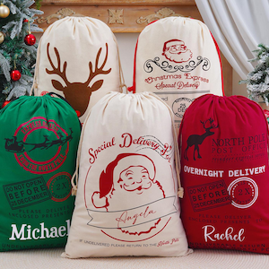 Op de afbeelding: Een verzameling kerstcadeautassen met trekkoord. Ontwerpen omvatten een rendier, de kerstman en teksten zoals "Christmas Express" en "Overnight Delivery". Namen als "Michael" en "Rachel" zijn ook te zien.